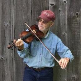 Man wearing red ball cap and denim button up shirt playing fiddle in front of barn